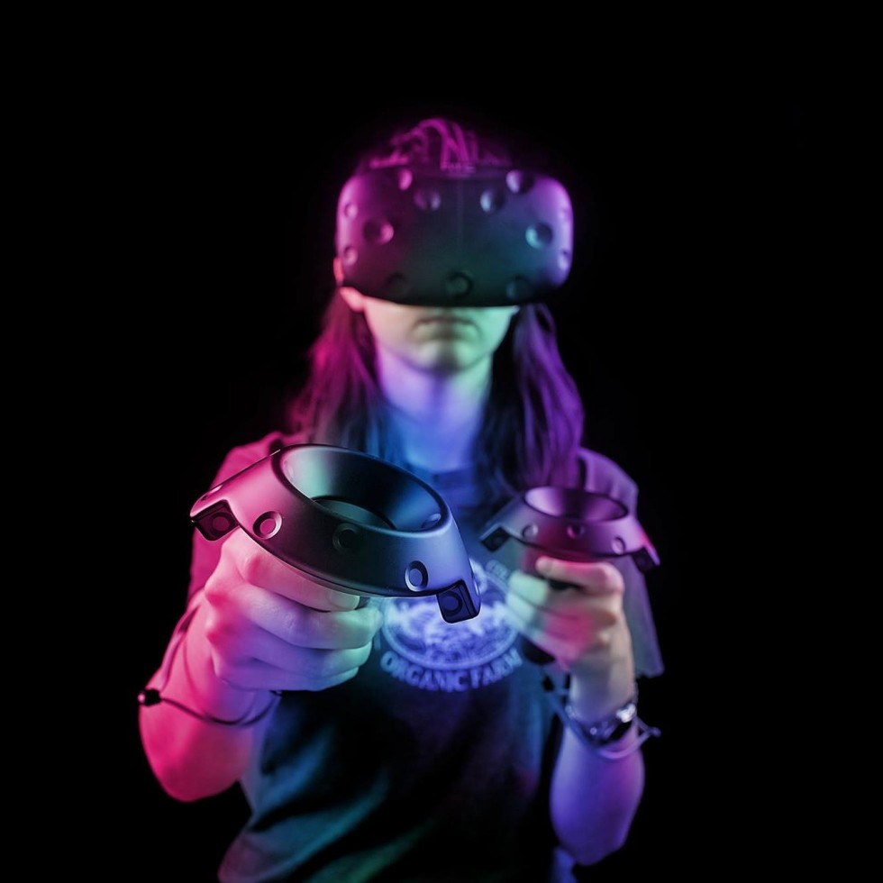 Miriam wears a virtual reality headset, one hand is in focus holding the device's vr remote toward the viewer. The lighting is pink and purple.